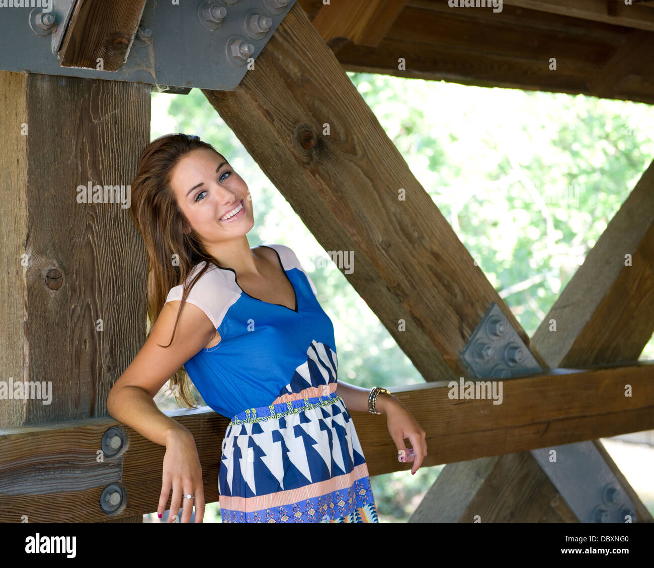 Attractive woman with long brown hair wearing a blue dress leans on the side of a covered wooden bridge. Stock Photo