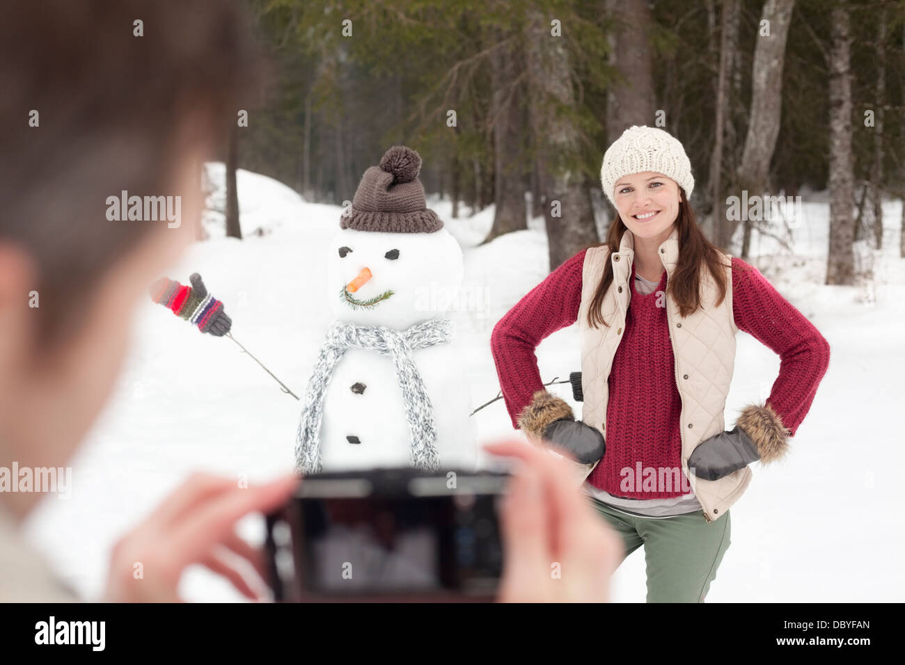 Man photographing smiling woman with hands on hips next to snowman in woods Stock Photo
