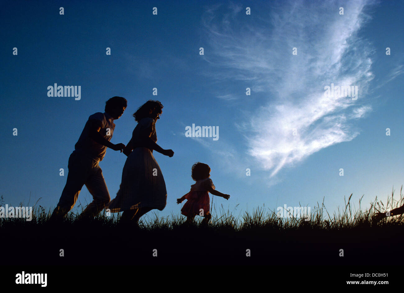 1980s SILHOUETTED FAMILY MOTHER FATHER CHILD GIRL RUNNING IN GRASSY FIELD Stock Photo