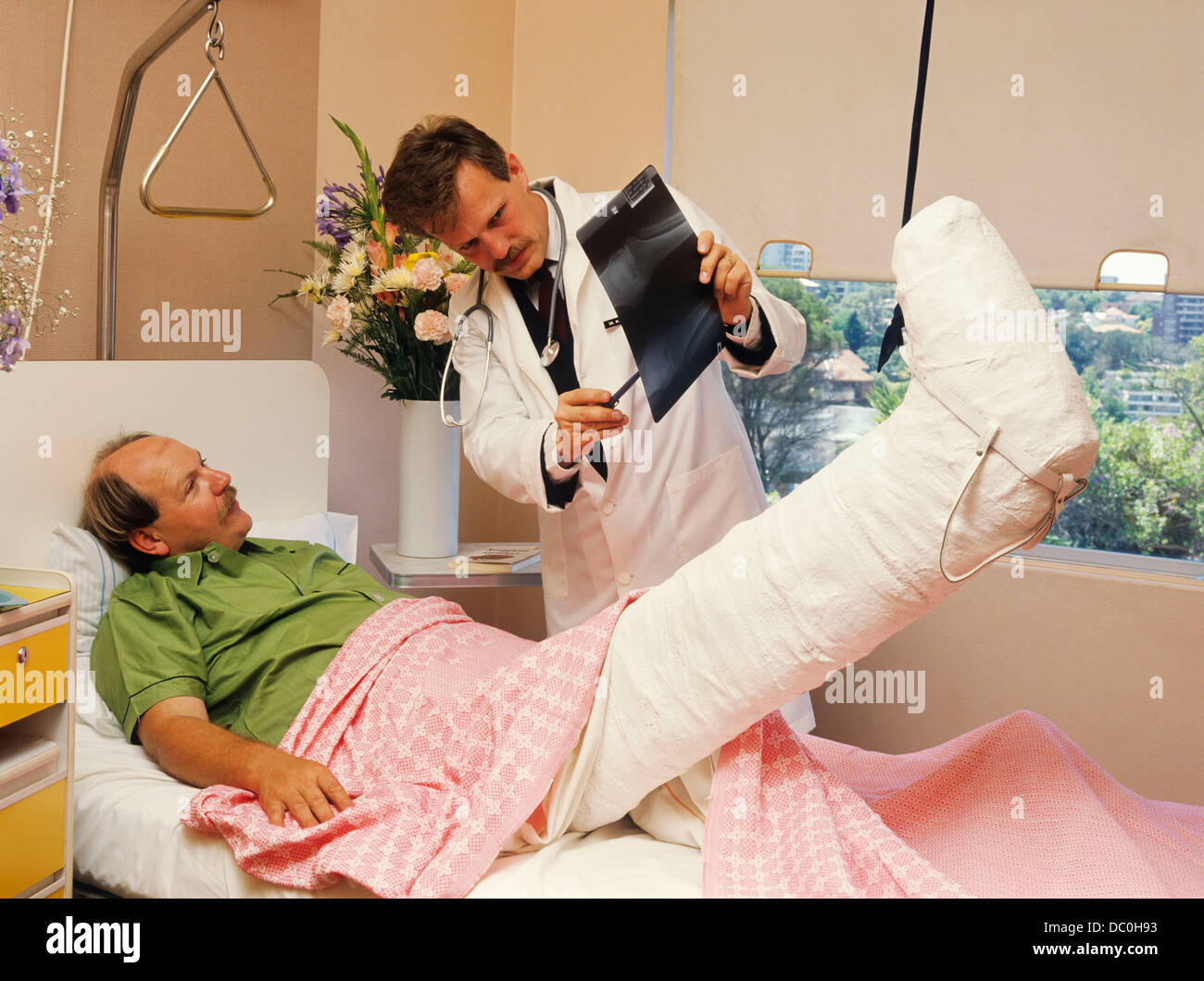 1980s 1990s MAN HOSPITAL BED BROKEN LEG IN CAST AND TRACTION GOING OVER X-RAY WITH DOCTOR Stock Photo