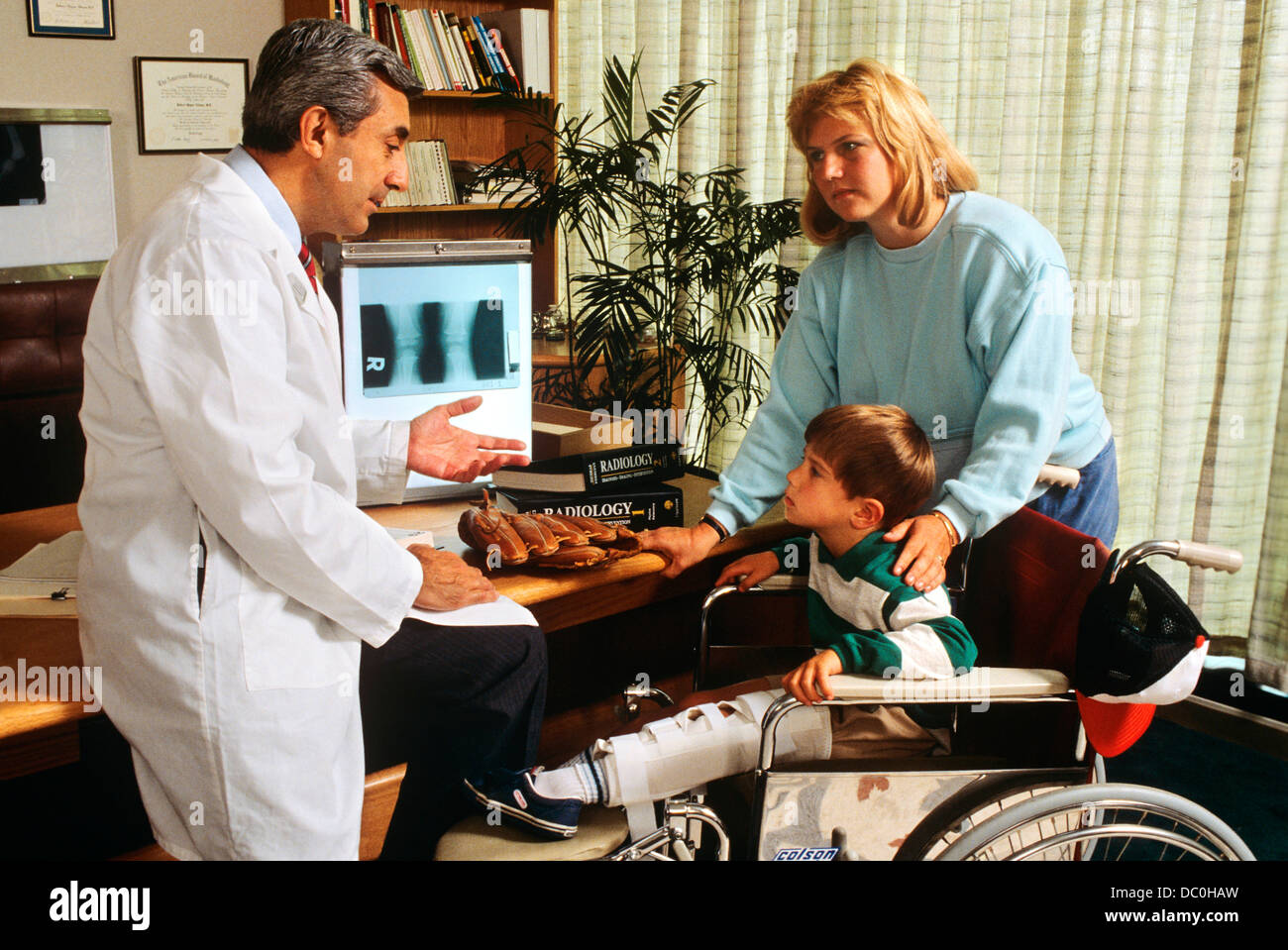 1990s DOCTOR EXPLAINING DIAGNOSIS TO SMALL BOY IN WHEELCHAIR AND HIS