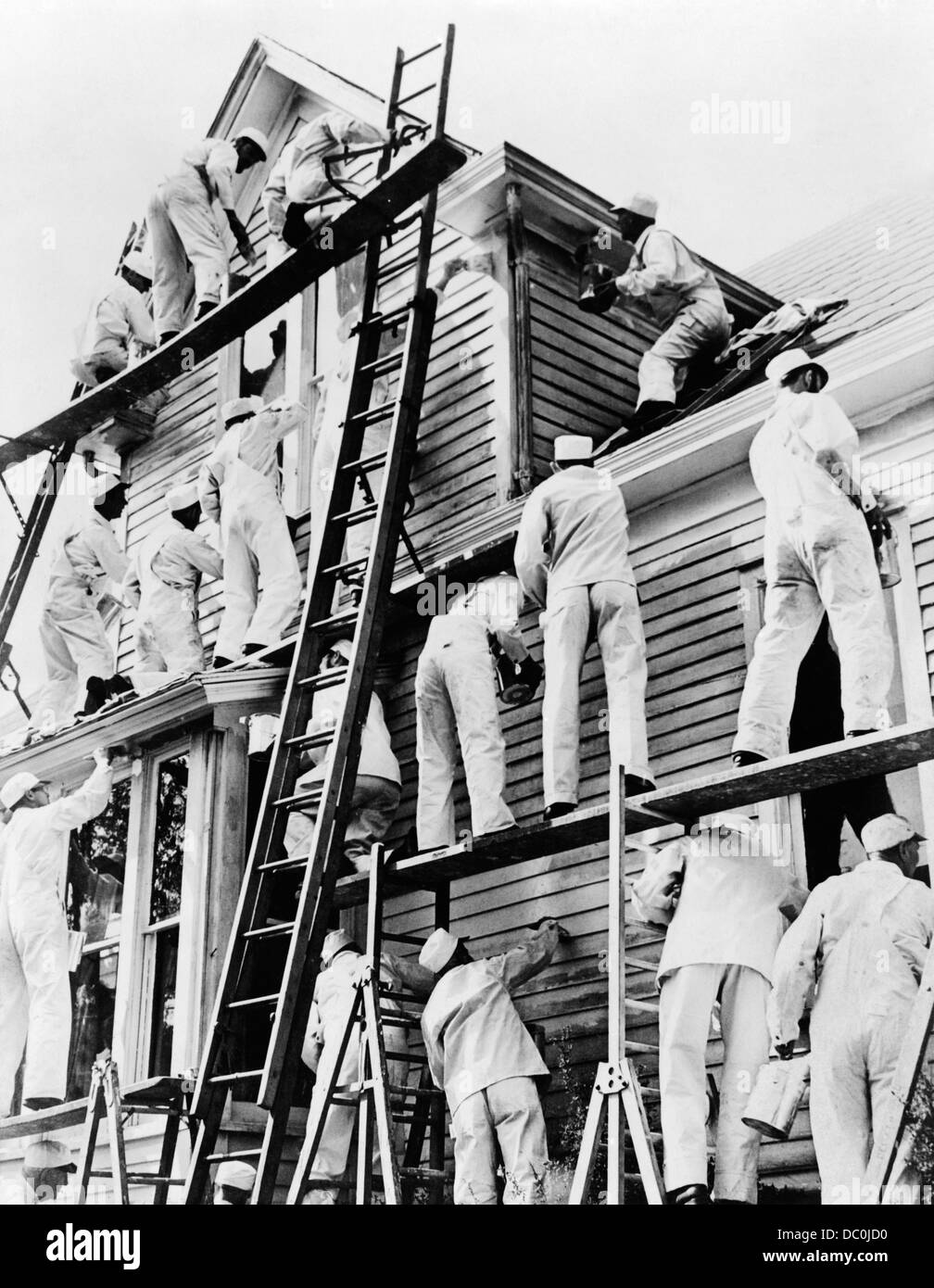 1930s GROUP 18 MEN PAINTERS IN WHITE WORK OVERALLS COVERALLS ON LADDERS AND SCAFFOLDS PAINTING A CLAPBOARD WOOD FRAME HOUSE Stock Photo