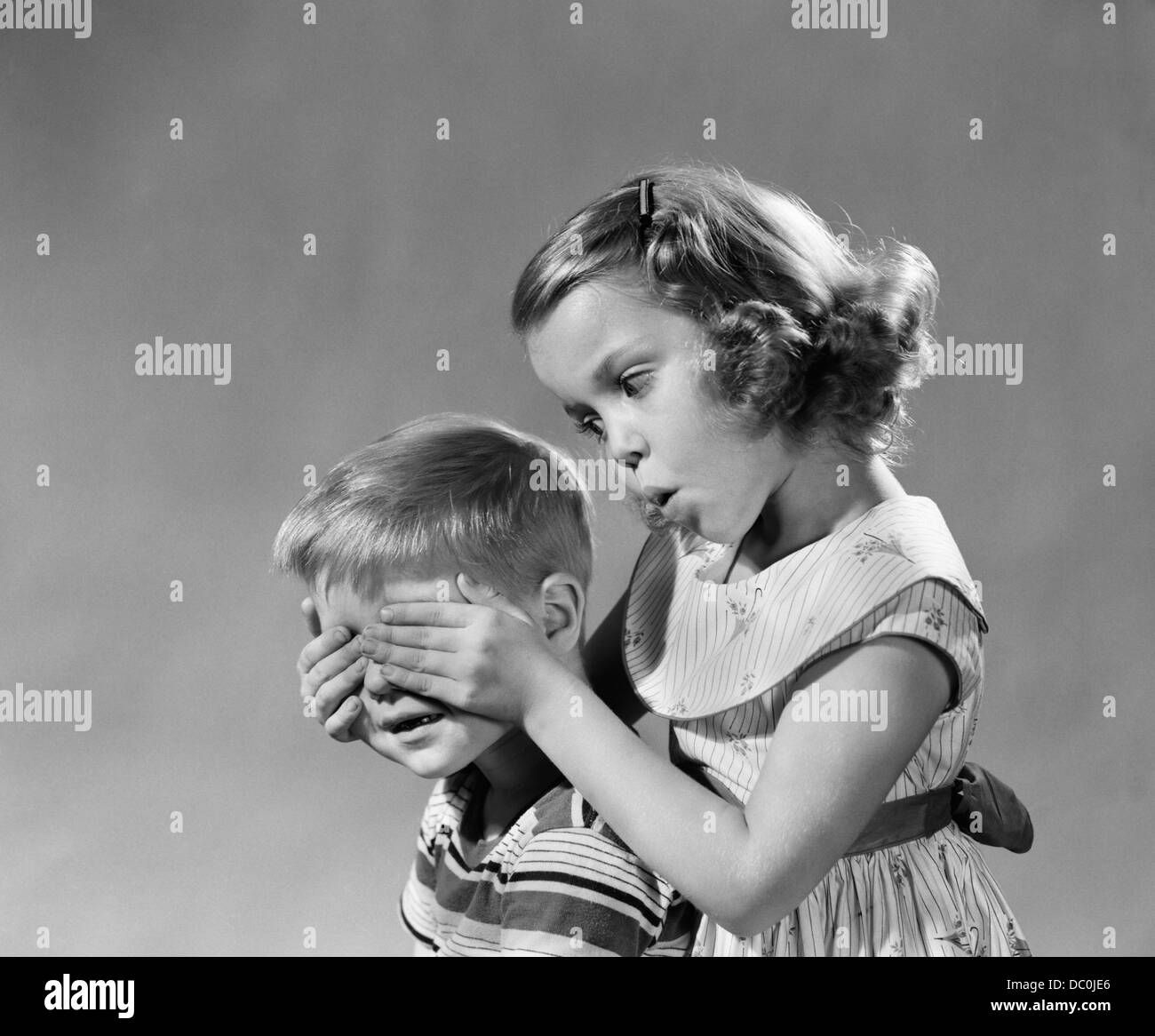 1950s 1960s GUESS WHO GIRL HOLDS HER HANDS OVER BOYS EYES BLIND GAME TEASING PLAYING BOY GIRLS Stock Photo