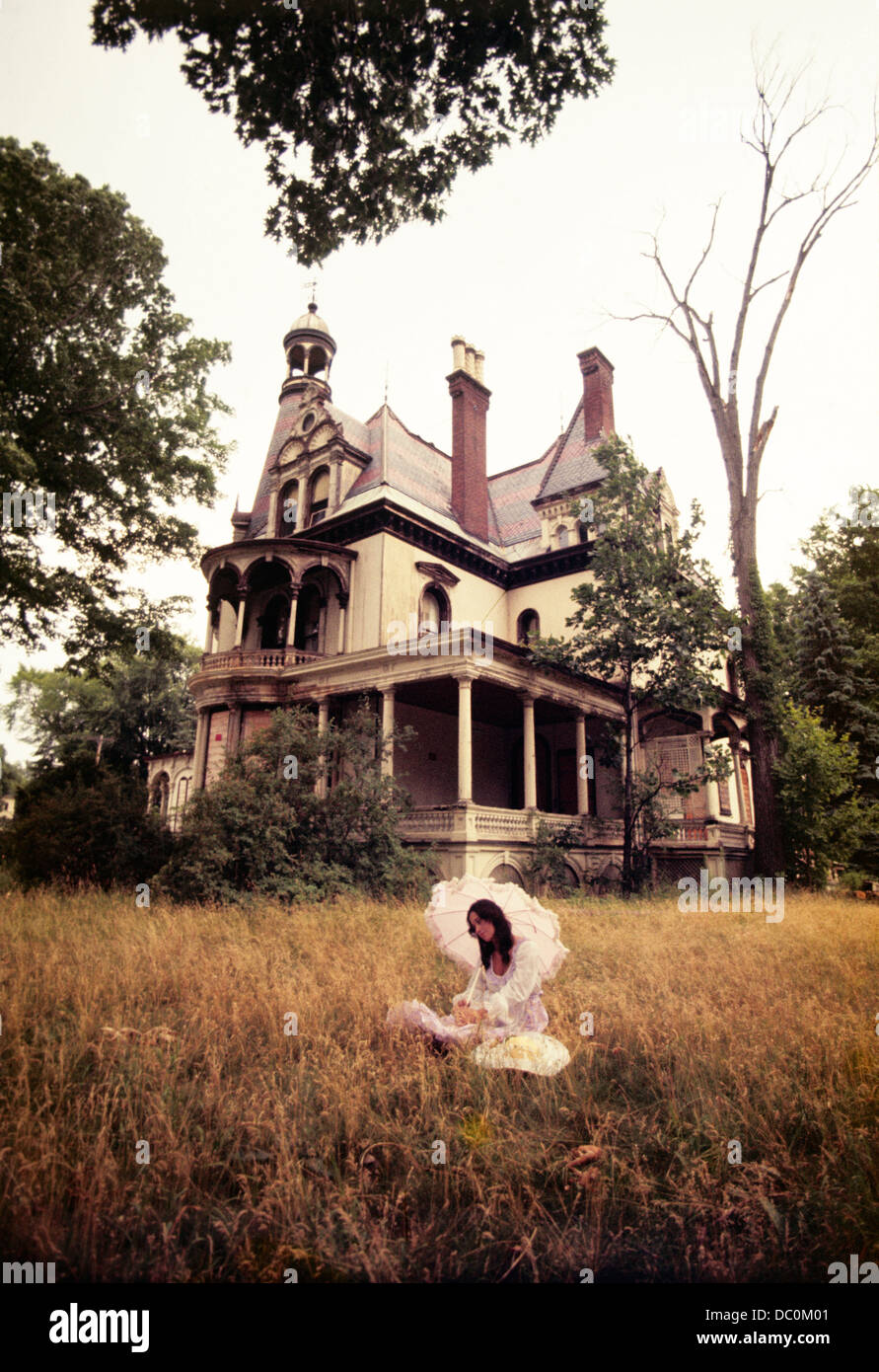 1960s 1970s WOMAN UNDER PARASOL SITTING FRONT OF ABANDONED HAUNTED VICTORIAN HOUSE Stock Photo