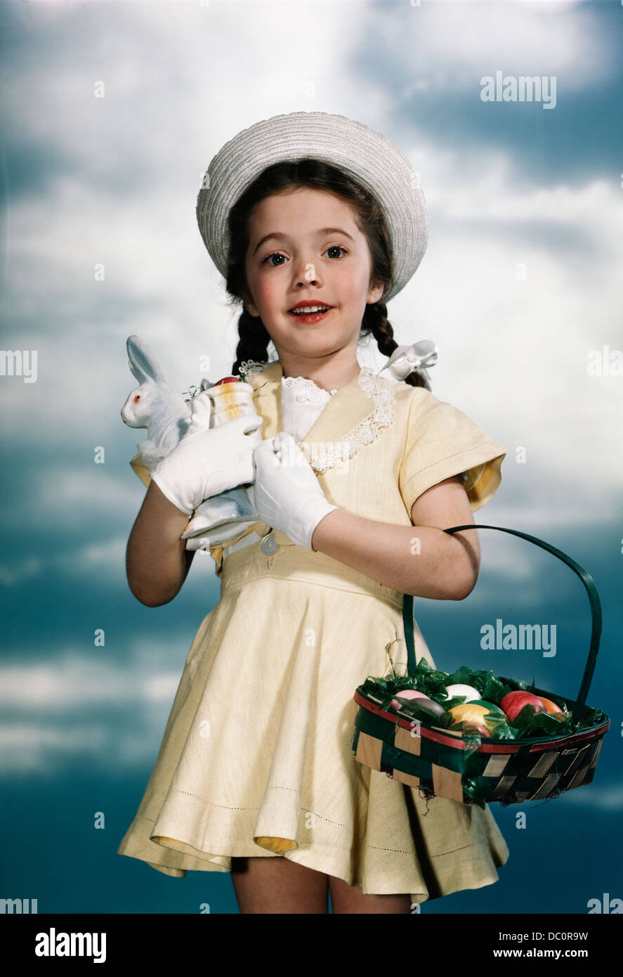 1950s SMILING GIRL WEARING YELLOW DRESS STRAW HAT HOLDING TOY RABBIT AND EASTER BASKET LOOKING AT CAMERA Stock Photo