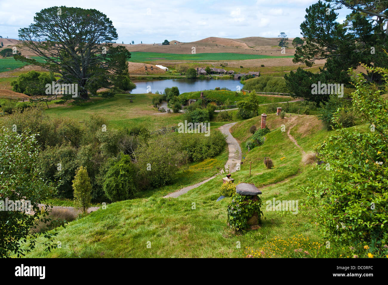 Very green Hobbiton with meandering path through image with lake and Party tree in background, showing hobbit hole chimneys Stock Photo
