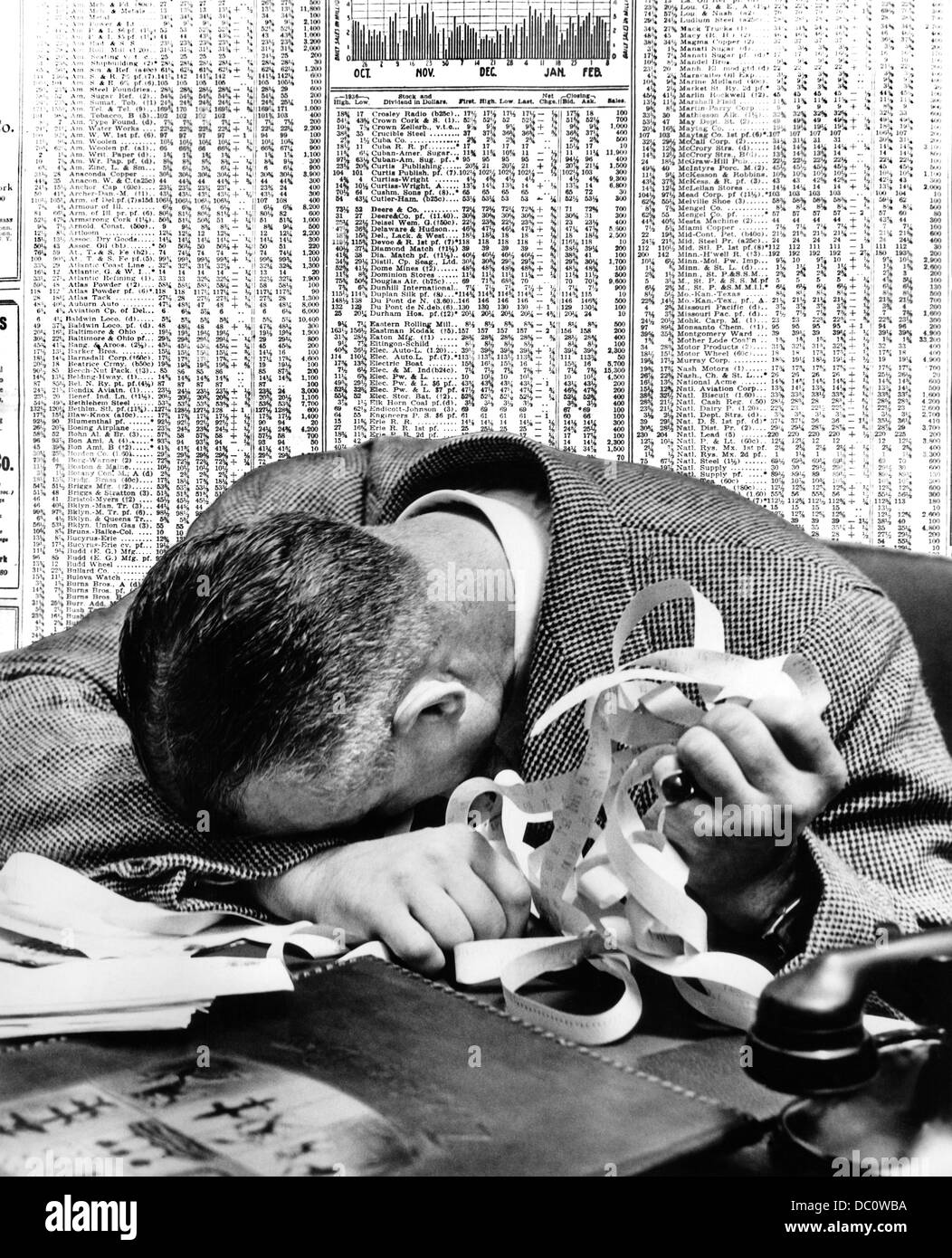 1940s MAN HEAD ON DESK HOLDING STOCK TICKER TAPE WITH NEWSPAPER STOCK PAGE BEHIND HIM Stock Photo