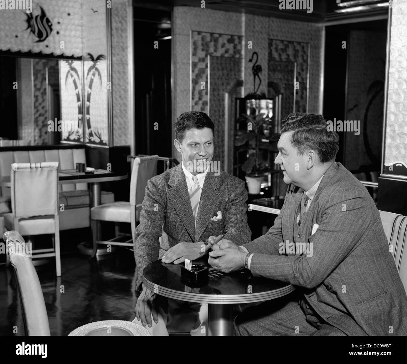 1930s 1940s TWO SMILING MEN FRIENDS SITTING TOGETHER AT TABLE IN NIGHTCLUB TALKING SMOKING CIGAR Stock Photo