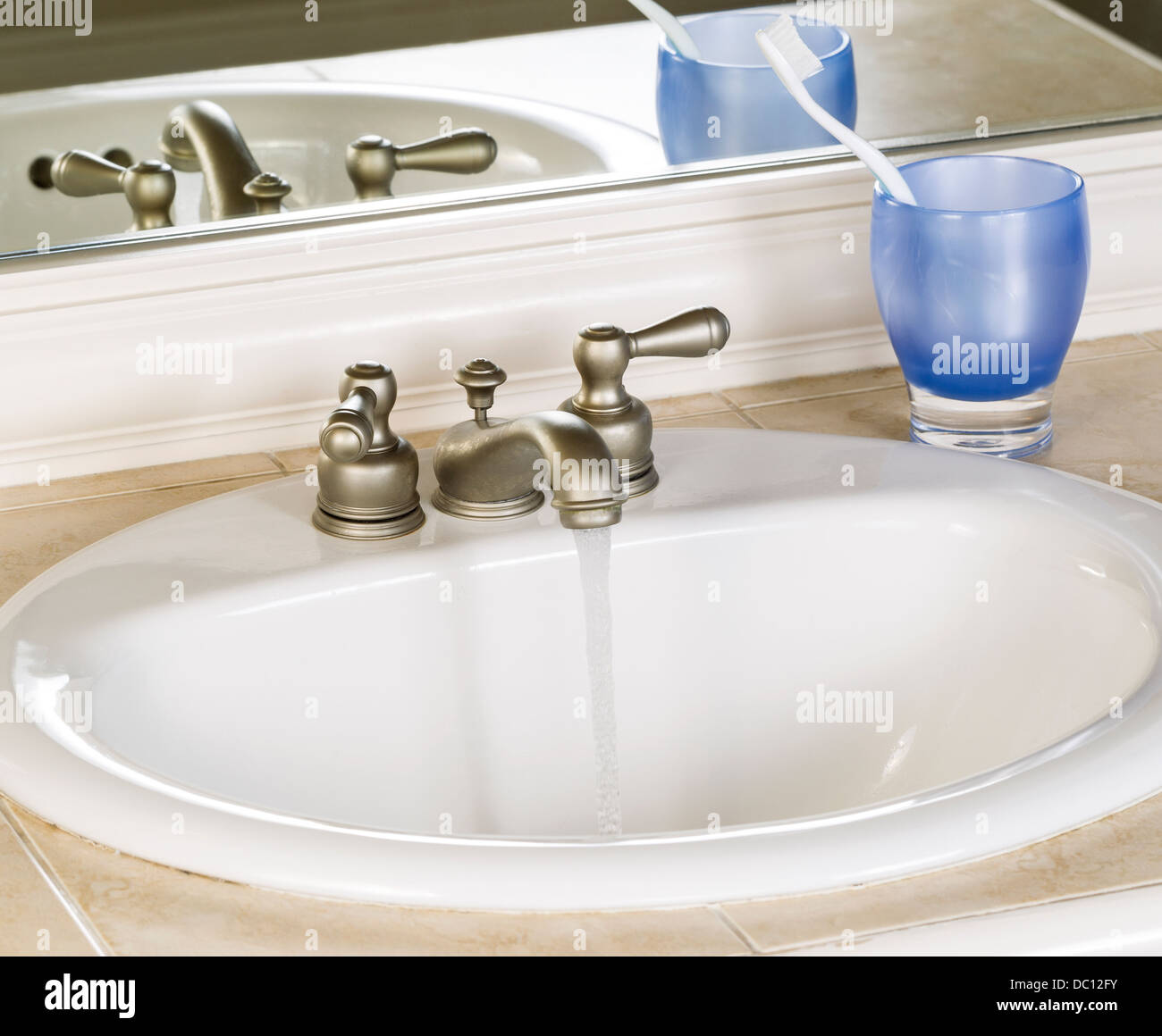 Photo of white bathroom sink and running faucet with blue cup, tooth brush and mirror in background Stock Photo