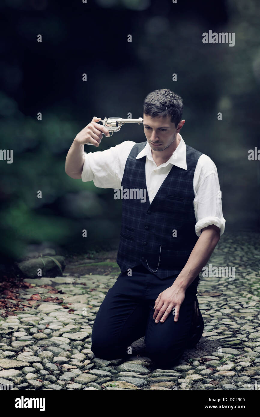 a man in vintage clothes is sitting on the ground, trying to shoot himself Stock Photo