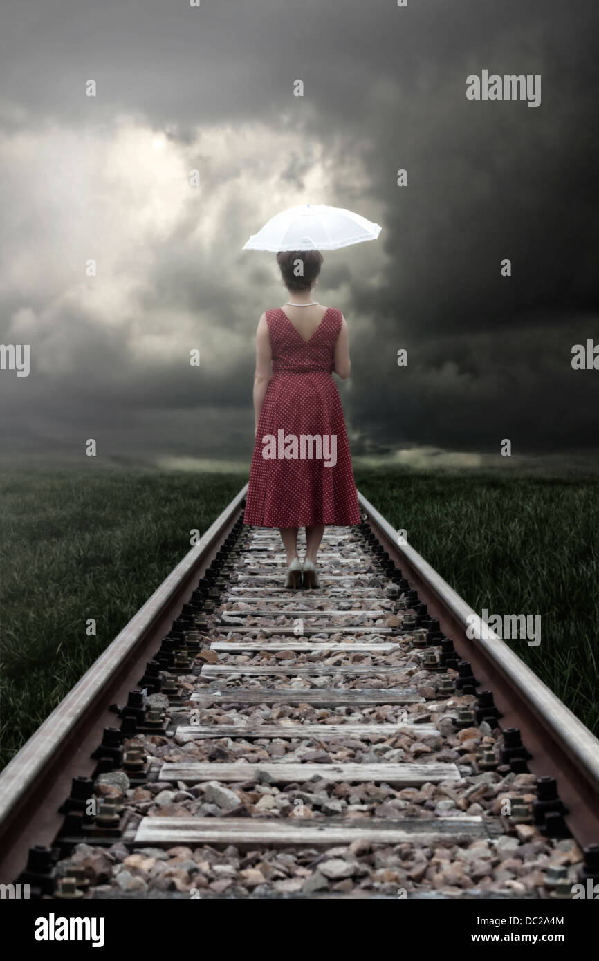 a girl in a red dress with a parasol is standing on railway tracks Stock Photo