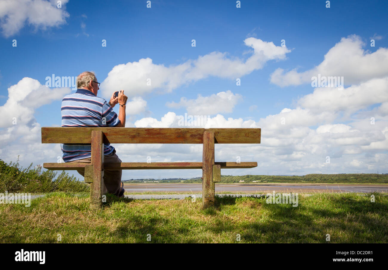 Elderley man sat on bench taking photographs at River Lune, Glasson Dock, Lancaster district, Lancashire Stock Photo