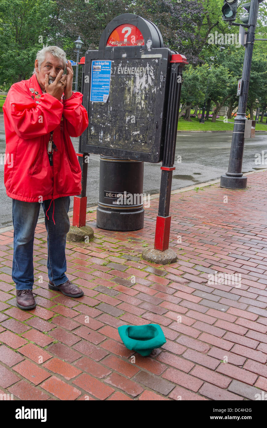 An older adult beggar male plays the harmonica outside on the sidewalk in a bright red coat, and gray hair. Stock Photo