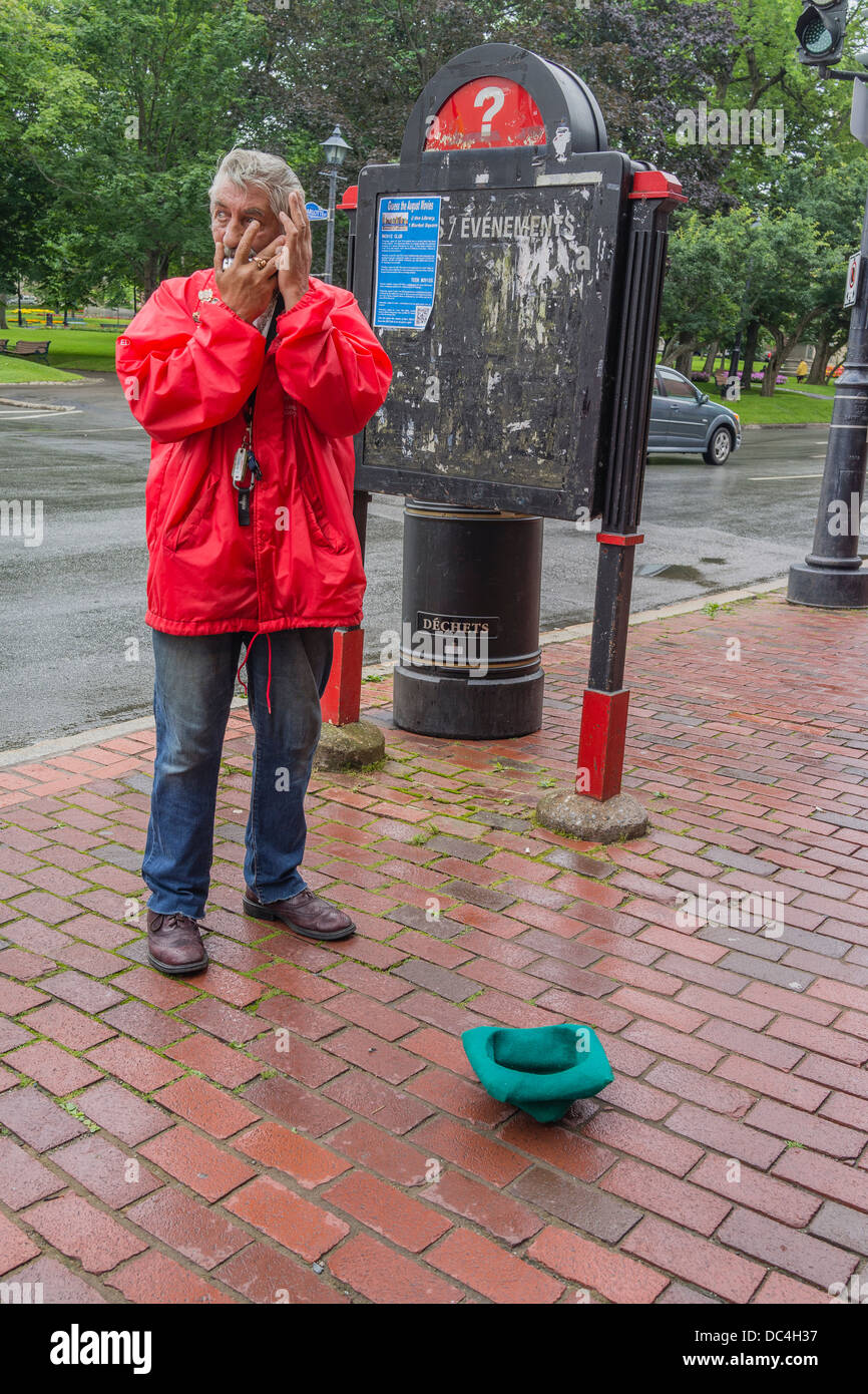 An older adult beggar male plays the harmonica outside on the sidewalk in a bright red coat, and gray hair. Stock Photo