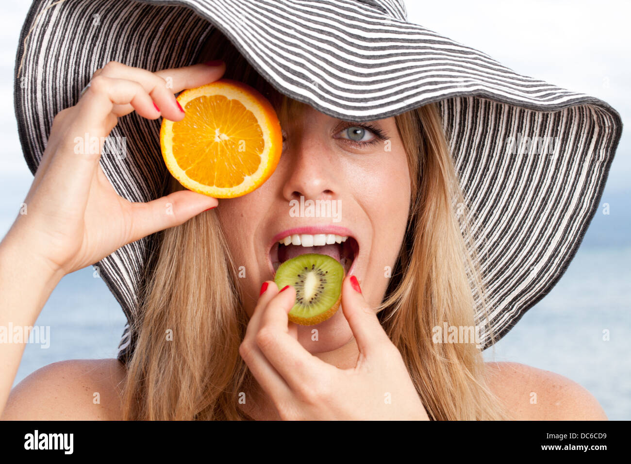 A woman covers one eye with an orange slice while taking a bite of kiwi. Stock Photo