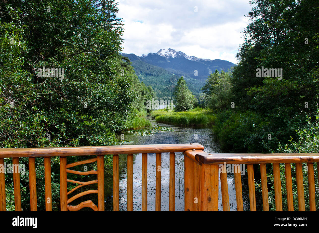 Ornate wooden bridge over small river, with mountains of Whistler in the distance.  In summer.  In Whistler, British Columbia,CA Stock Photo