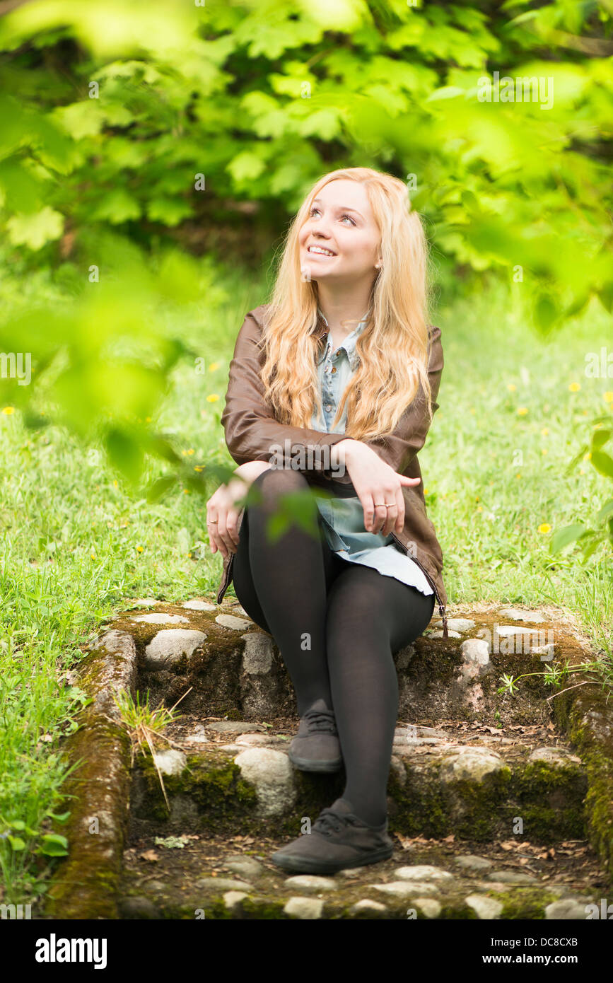 Nature scene with one young attractive blonde woman sitting in a park looking up Stock Photo