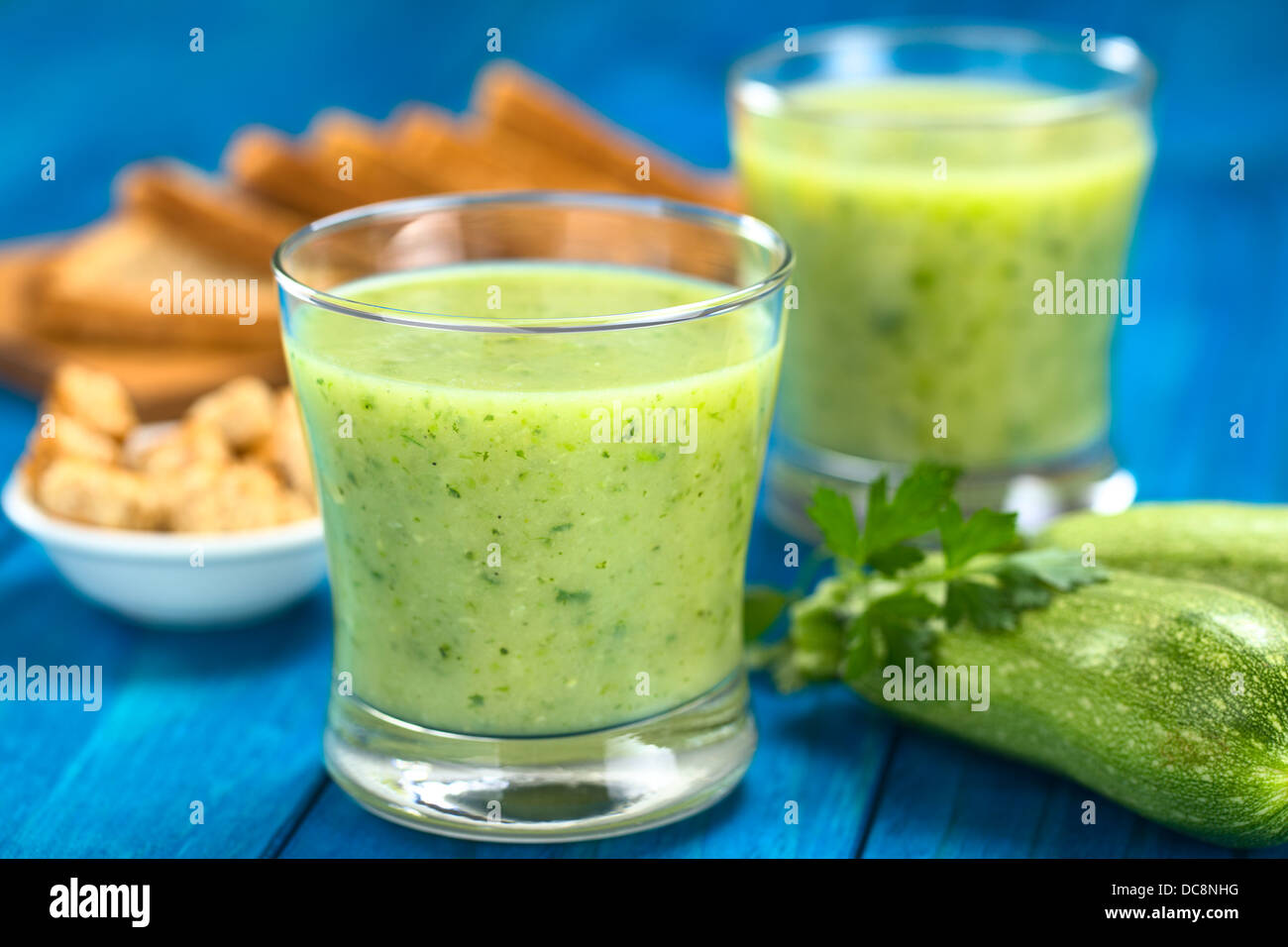Zucchini cream soup served in glasses on blue wood with small zucchini and parsley on the side, croutons and toast bread Stock Photo