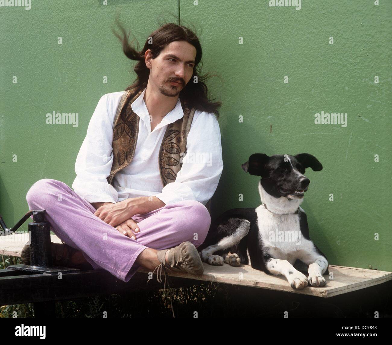 A Long Haired Young Man In Hippie Clothes With His Dog