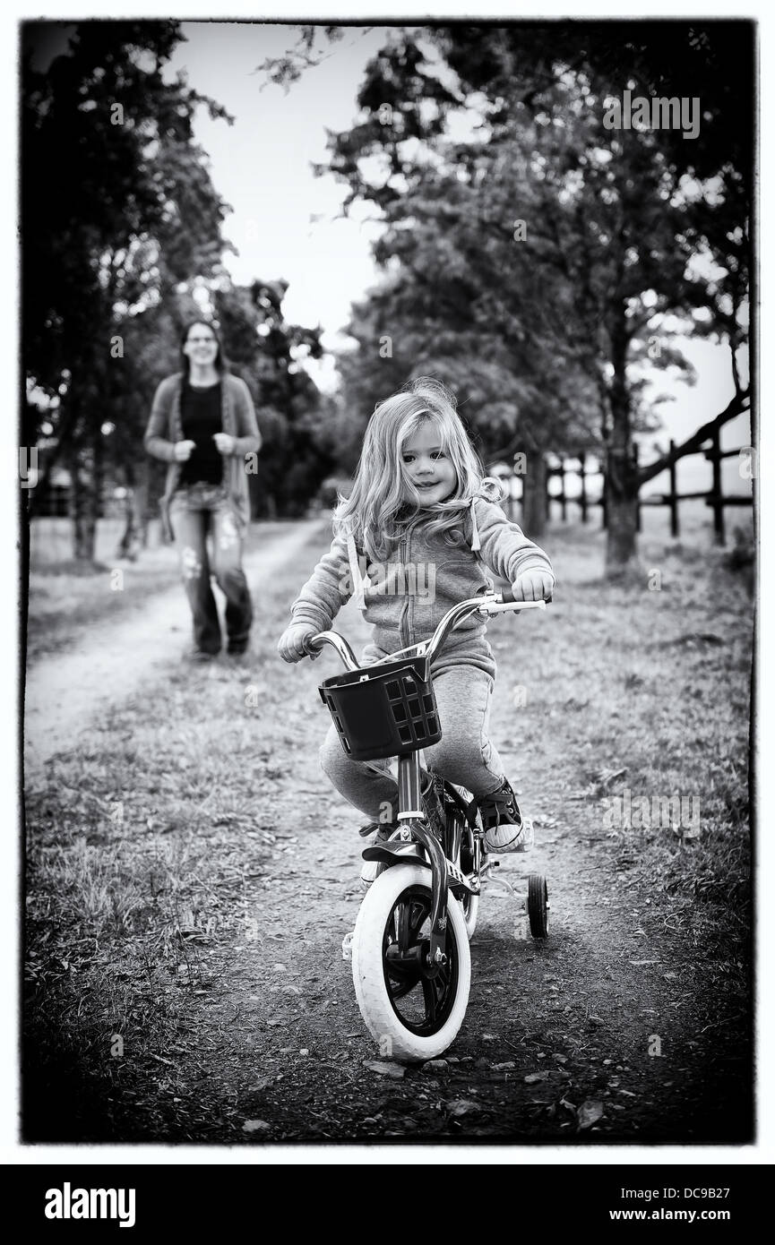 Cute toddler girl on her 1st bicycle with her mom running behind her. Also available in a black & white photo. Stock Photo
