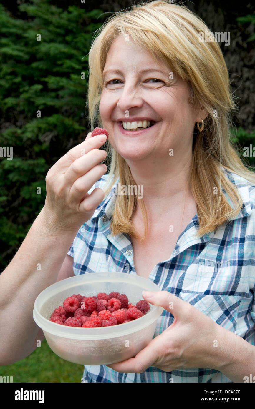 Smiling Caucasian woman eating freshly picked home grown raspberries, from garden in Bristol, UK Stock Photo