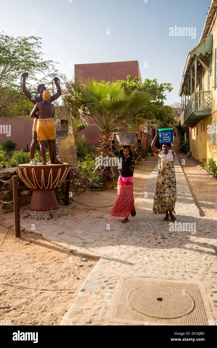 Women carrying Water Past Statue Commemorating the End of Slavery, Goree Island, Senegal. Stock Photo