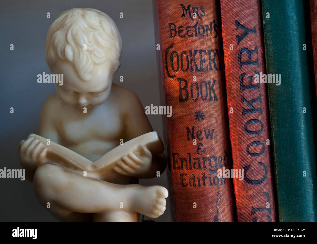 Carved bookend statue of boy reading alongside antique old cookery books in library Stock Photo