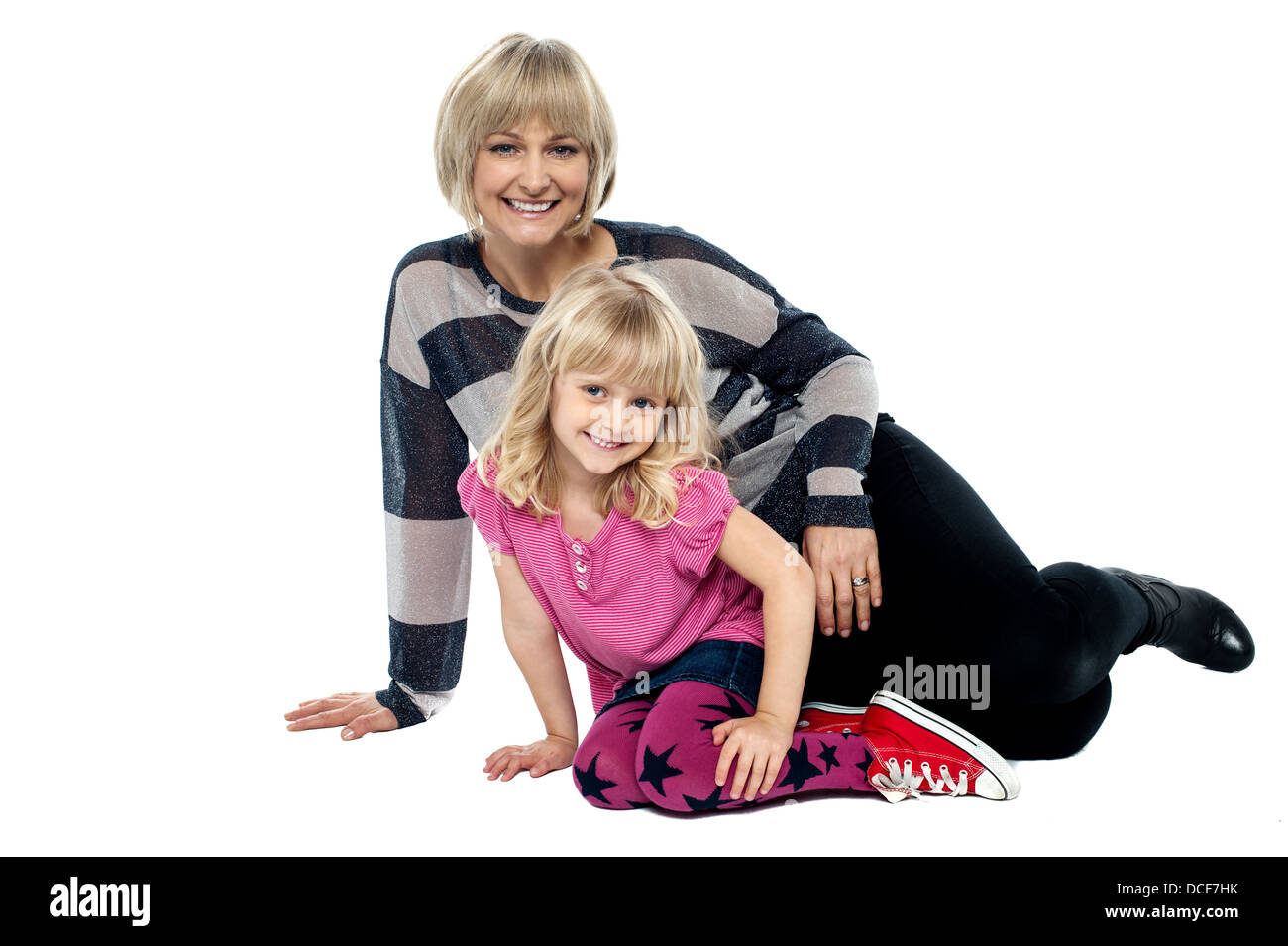 Cheerful mum and daughter sitting on studio floor. Facing camera and flashing smile. Stock Photo