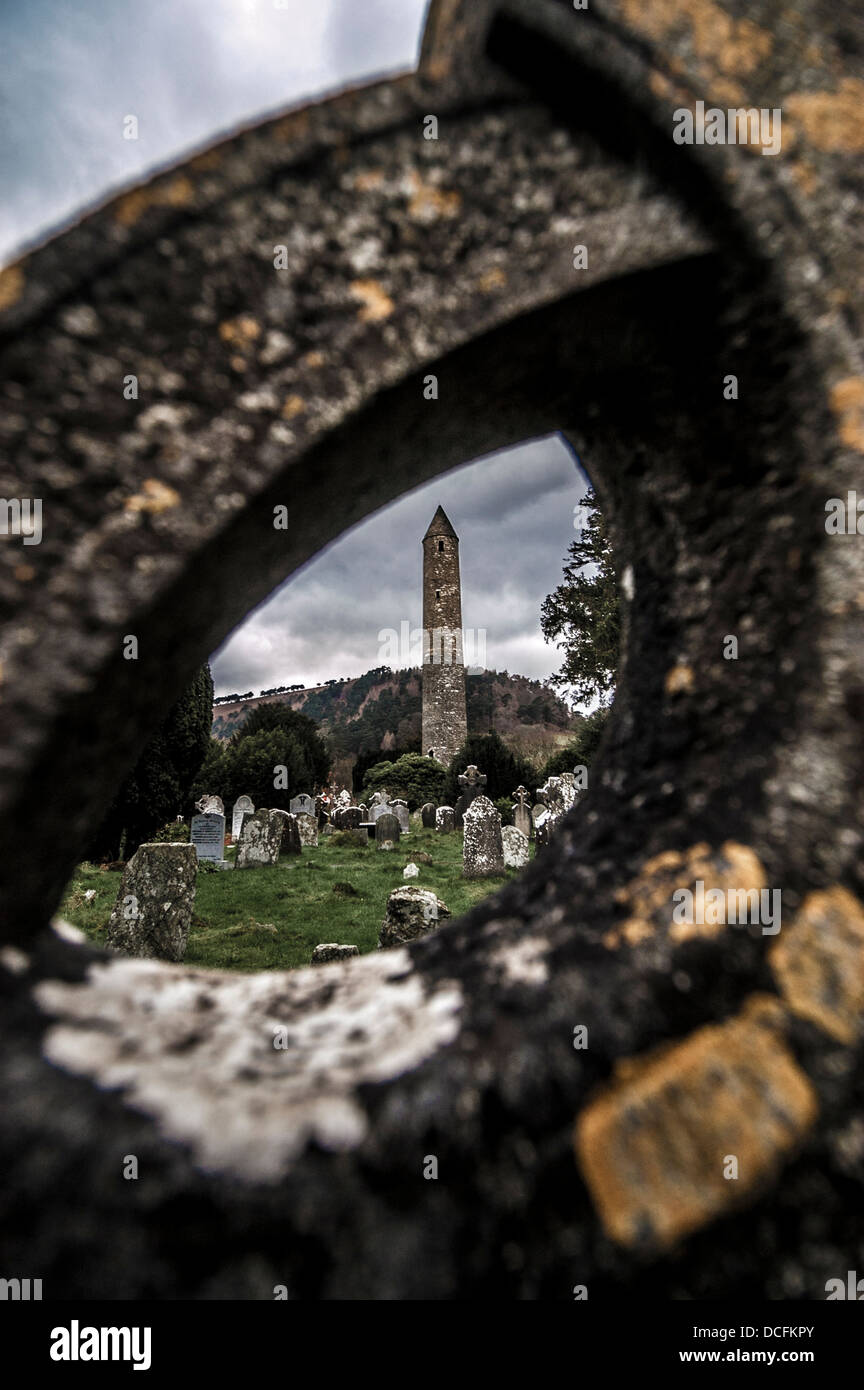 The Round Tower in the Monastic City, shot through a Celtic cross gravestone. Glendalough Valley, Ireland. Stock Photo