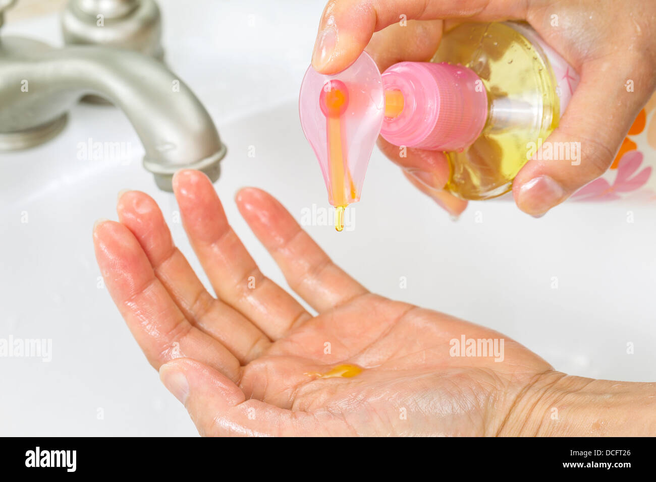 Horizontal photo of female hands putting liquid wash soap, from bottle, into hand with bathroom sink and faucet in background Stock Photo