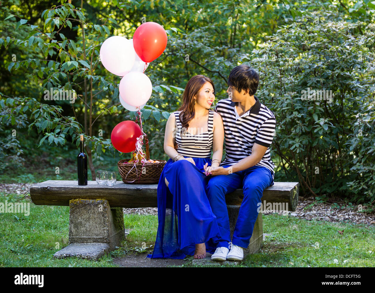 Horizontal photo of young adult couple, sitting on log bench, with balloons, basket of fruit, red wine, glasses and trees in bac Stock Photo