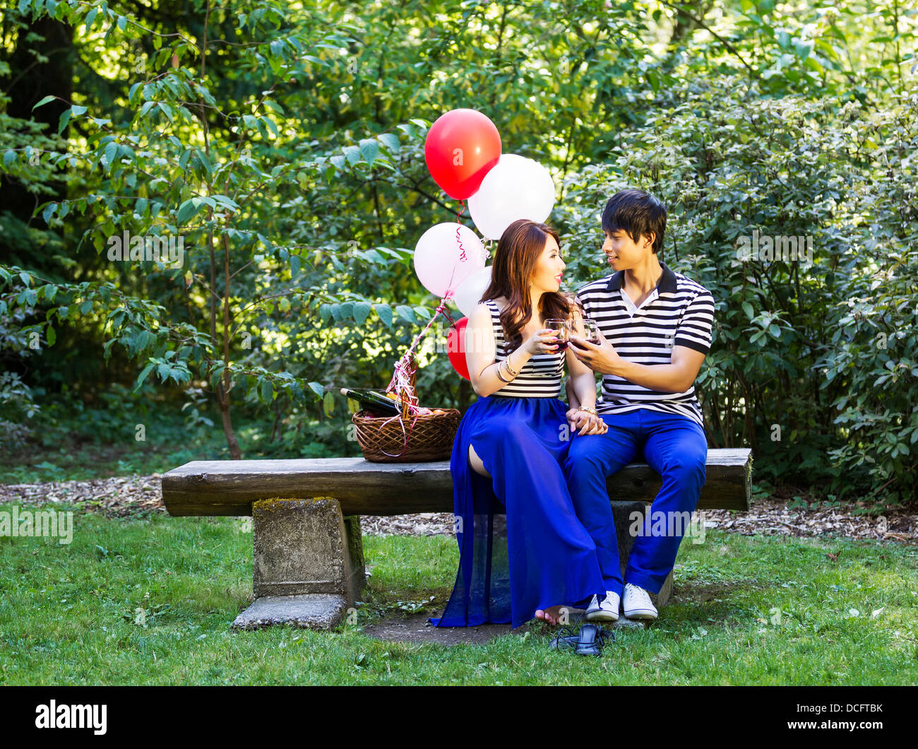 Horizontal photo of young adult couple sitting on log bench with glasses filled with red wine being held in their hands with bal Stock Photo