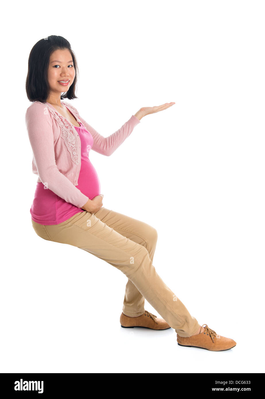 Full body six months pregnant Asian woman sitting on invisible chair, hand showing blank space looking at camera, isolated on white background. Stock Photo