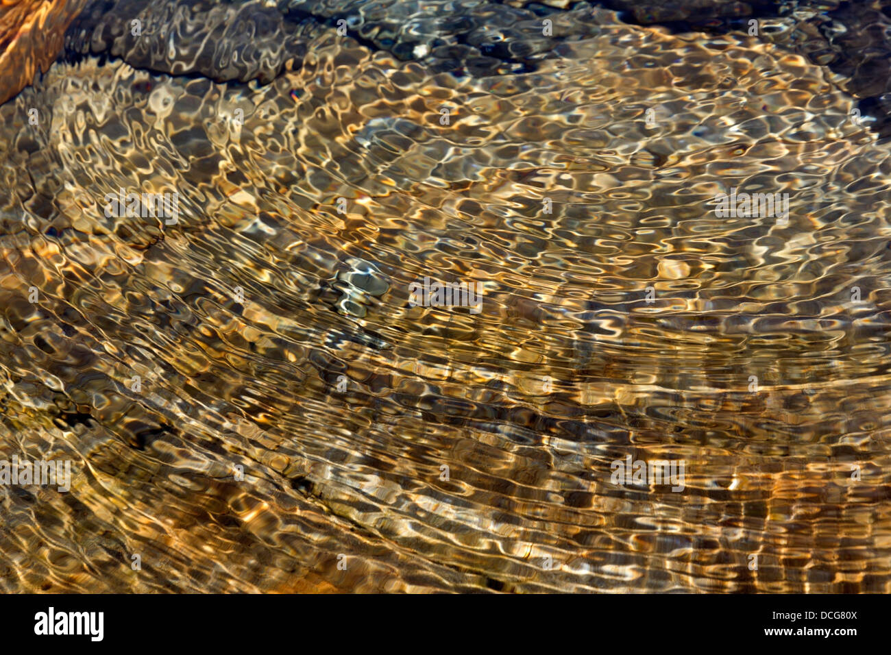 Colourful golden abstract circular water ripples above sunlit rocky stream bed, UK Stock Photo