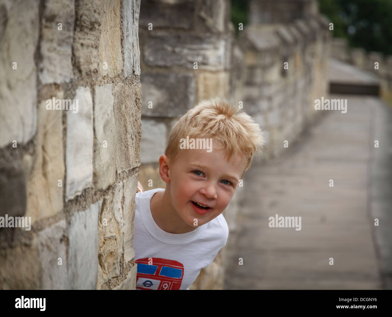 A little small blonde white Caucasian European British English boy peeping around a stone wall smiling laughing happy carefree Stock Photo