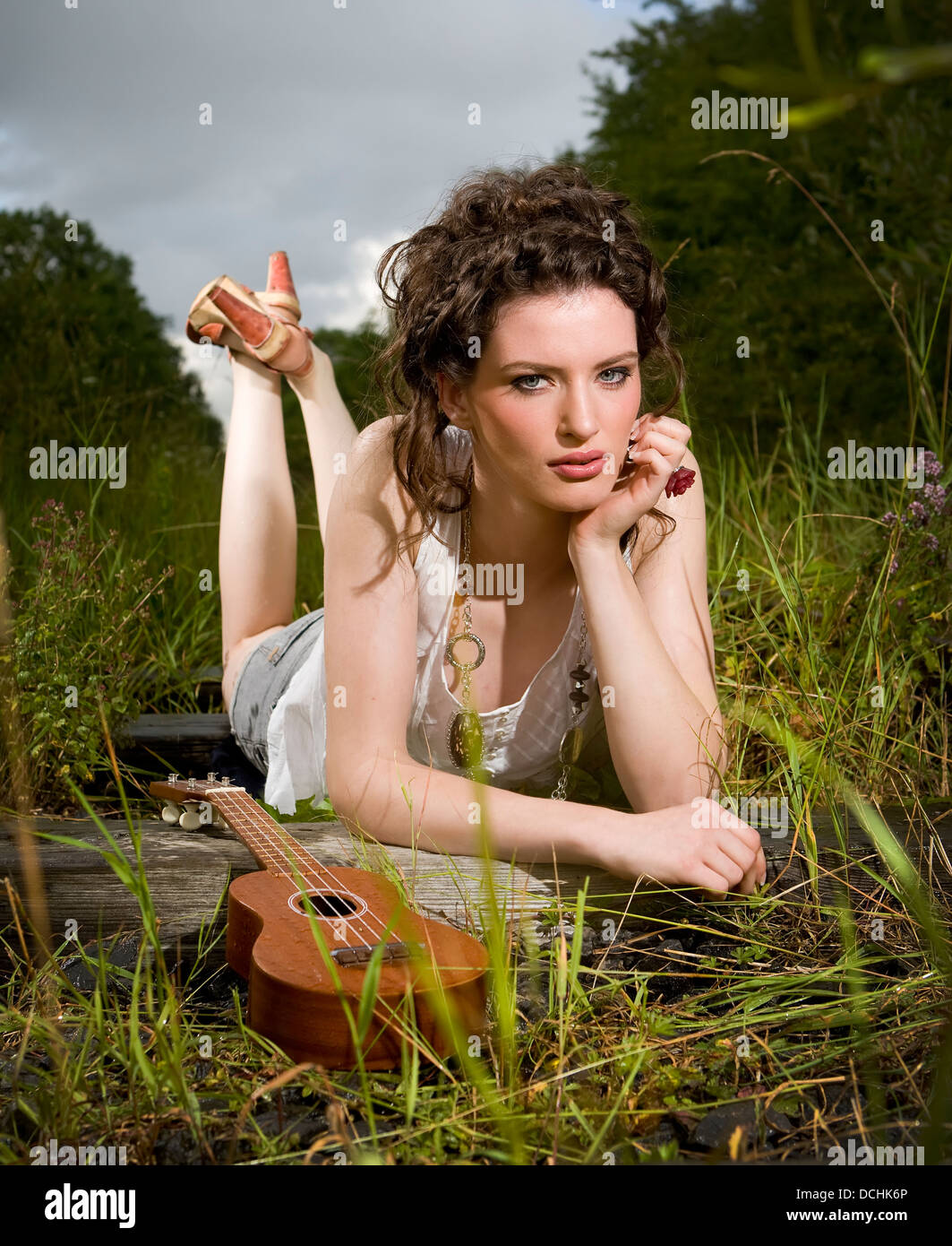 model with ukulele lying in grass looking into the camera on an old railway track wearing jewelery Stock Photo
