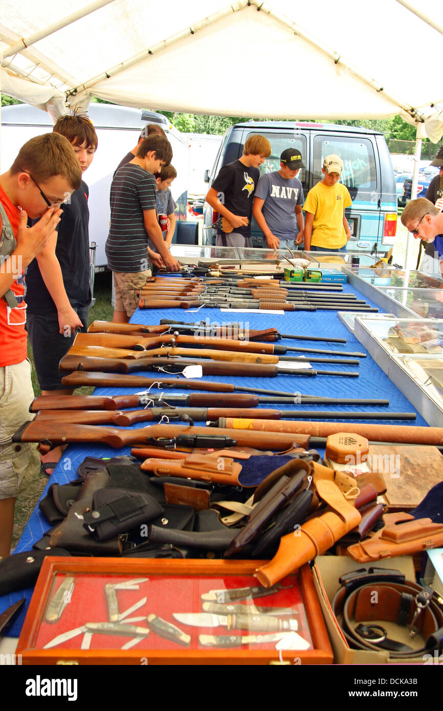 A group of young men gather around a table outside at a flea market, looking at guns, rifles, and other weapons for sale Stock Photo