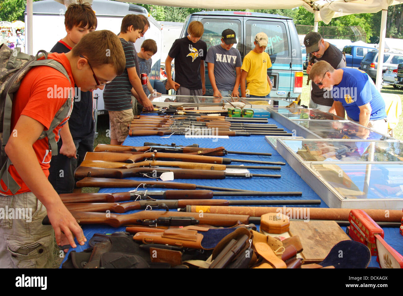 A group of young men gather around a table outside at a flea market, looking at guns, rifles, and other weapons for sale Stock Photo