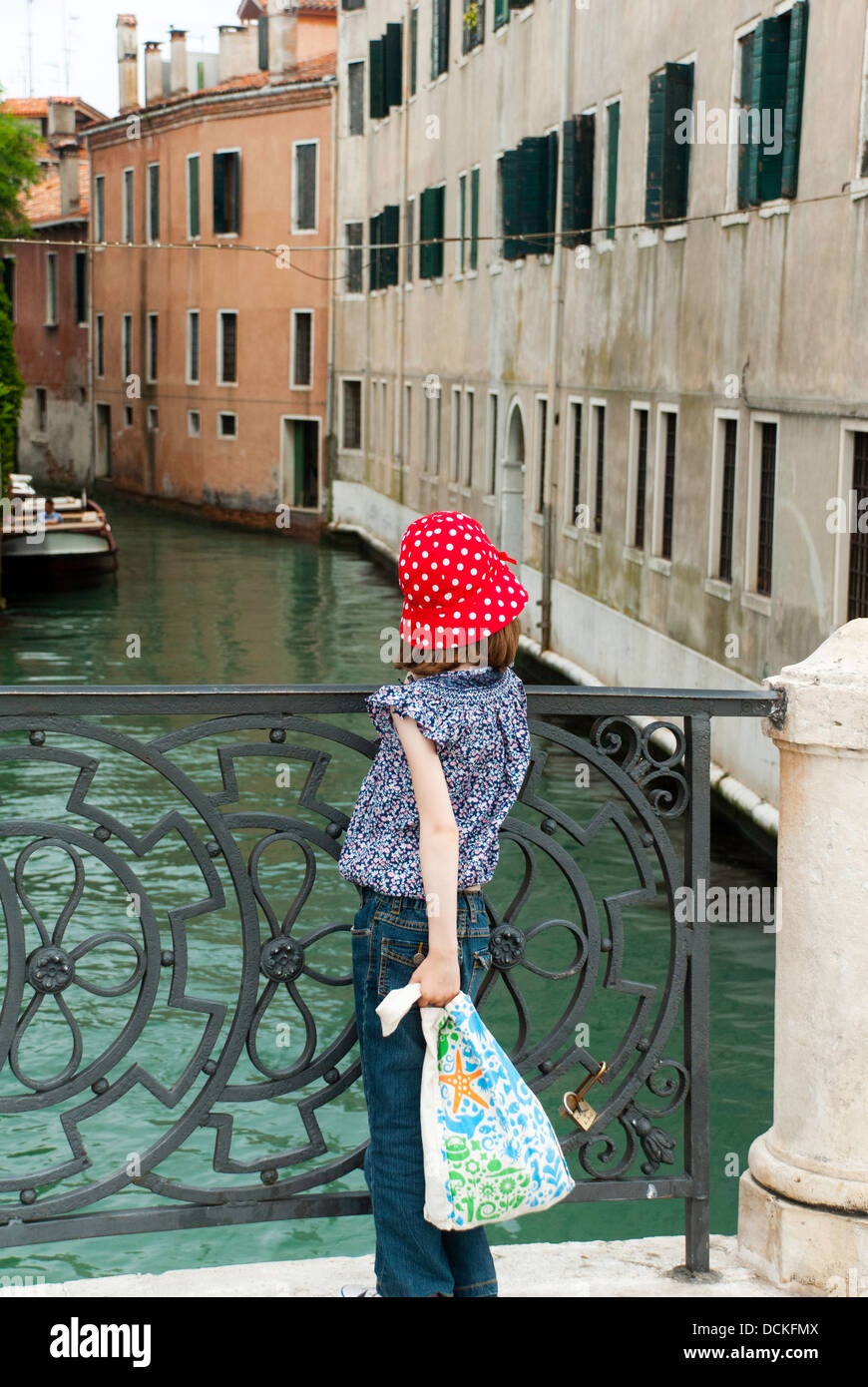 Young girl in red hat standing on a small bridge looking at a Venetian canal, Venice, Italy Stock Photo