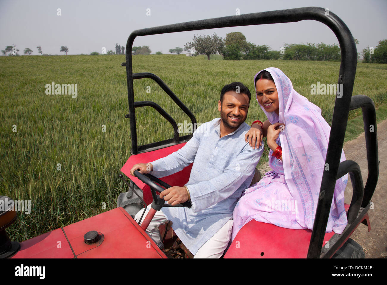 Portrait of an Indian couple sitting in a tractor with field in ...