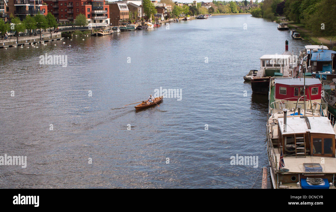 A lone man in a skiff makes his way down the River Thames in Kingston, London, England, past houseboats and flats. Stock Photo