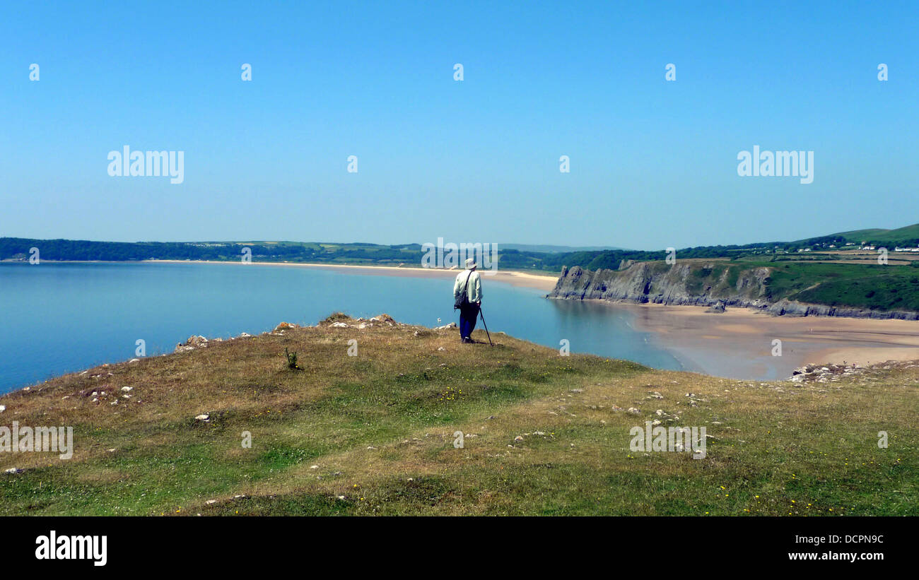 Single elderly figure with stick and sun hat wistfulon cliff edge overlooking Three Cliffs Bay on the Gower Peninsula, Swansea, South Wales, UK Stock Photo