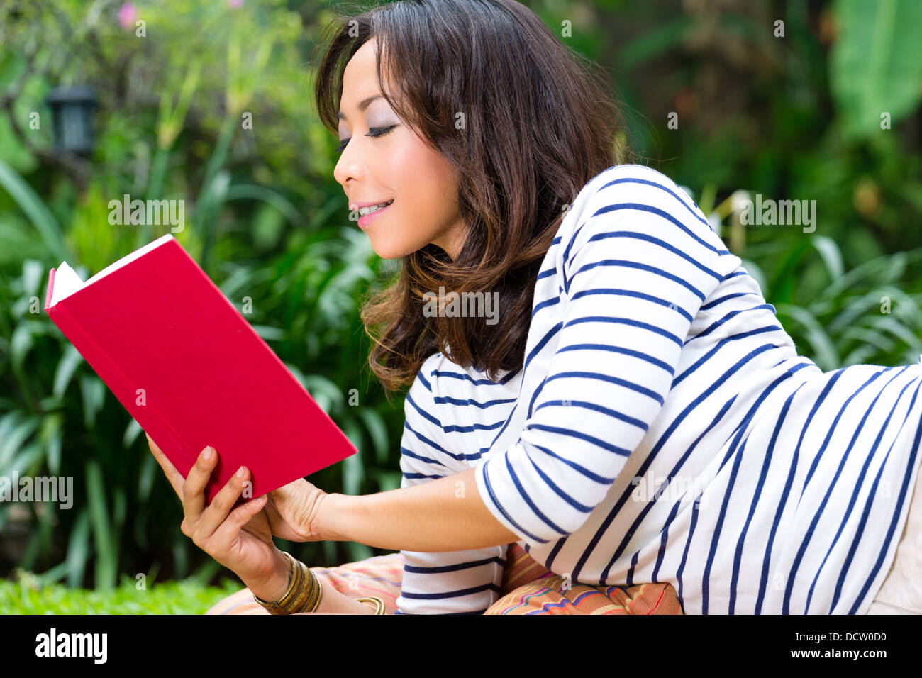 Young Indonesian woman lying at home in the garden and reading a book in her leisure time Stock Photo