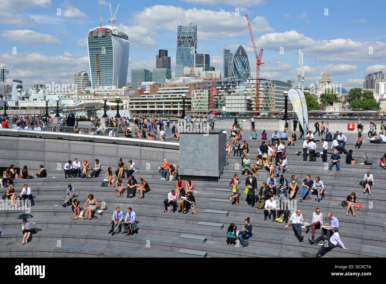 Groups of office workers summer lunch break time sitting on the steps of The Scoop with City of London skyscraper skyline beyond Southwark England UK Stock Photo