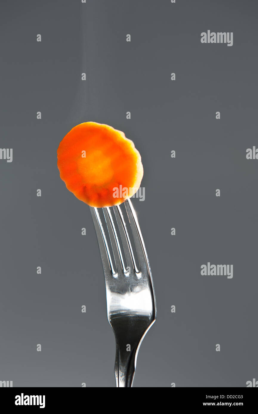 Hot round carrot slice on fork with steam on gray background. Stock Photo