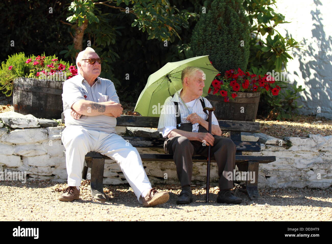 An old man uses an umbrella to shade from the sun while another man basks in the heat. Luss, Loch Lomond, Scotland, UK Stock Photo
