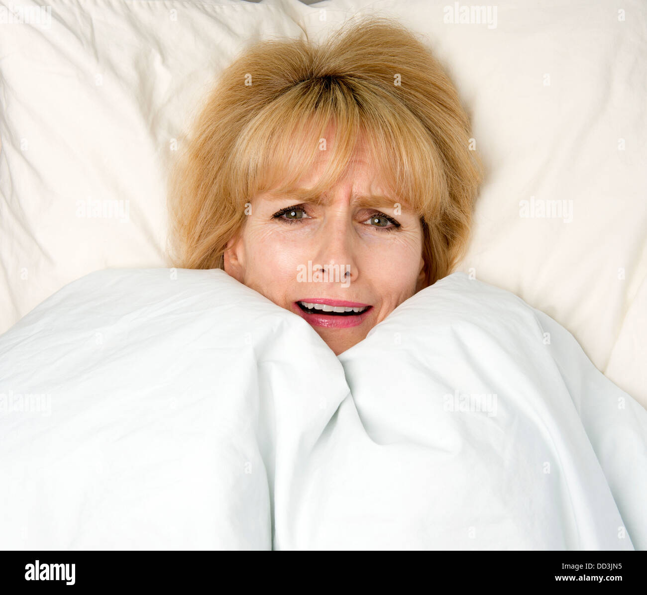 Woman pulling blanket to her face in bed with expression of fear Stock