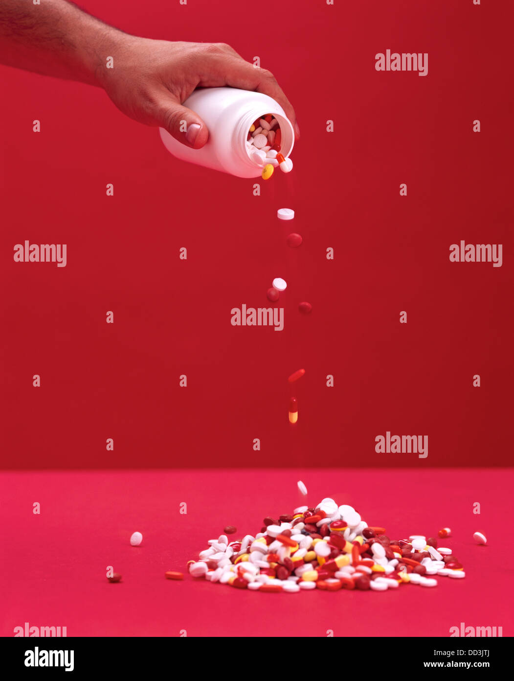 A hand pouring a container of medical pills into a large pile. Red background Stock Photo