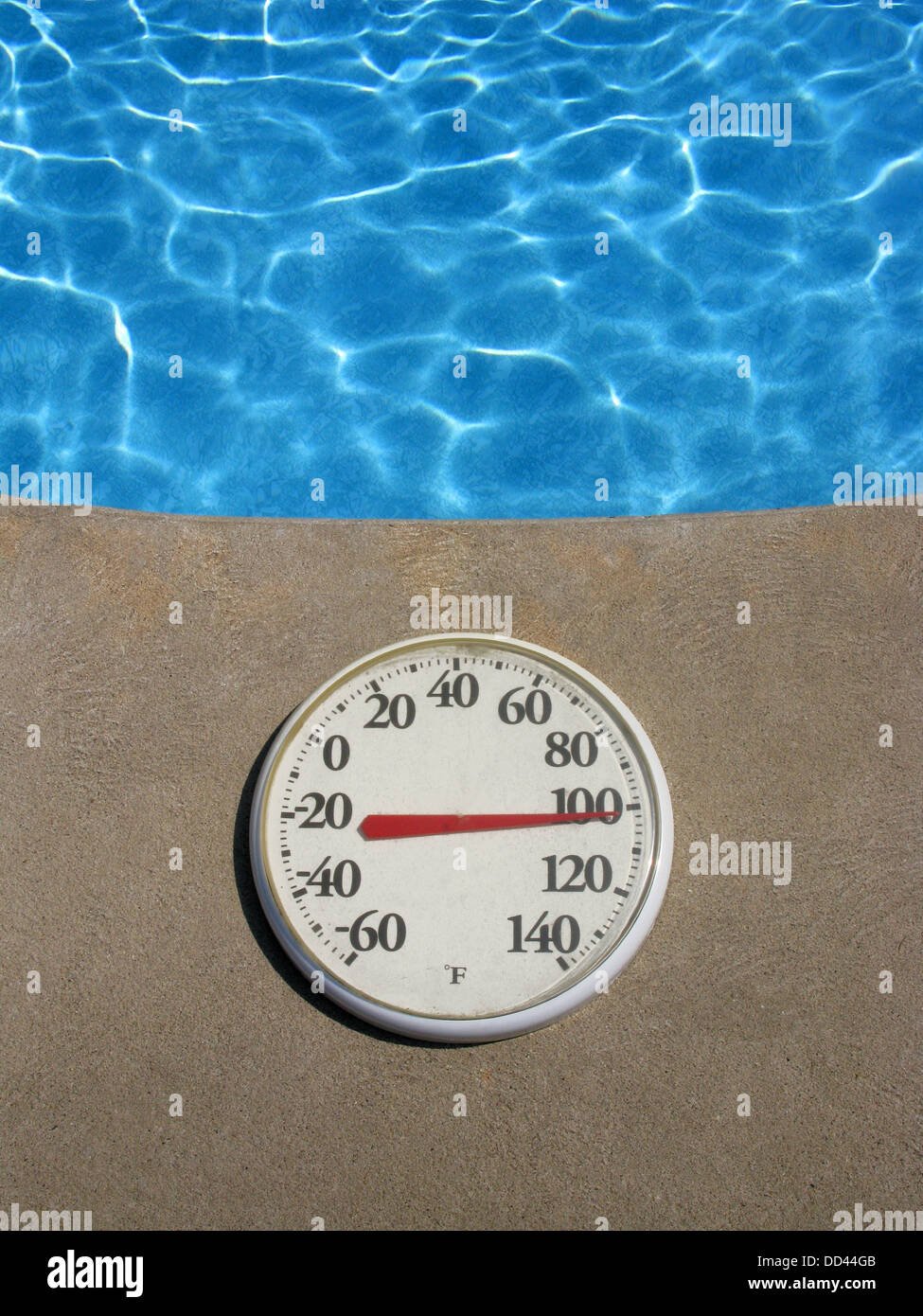 A round plastic thermometer showing hot temperature. Object is next to a swimming pool of blue water Stock Photo