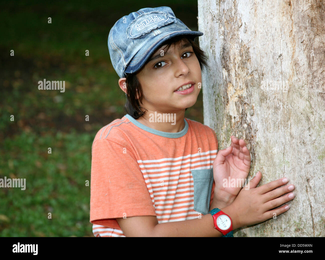 Portrait of brunette boy in baseball cap leaning on tree trunk Stock Photo